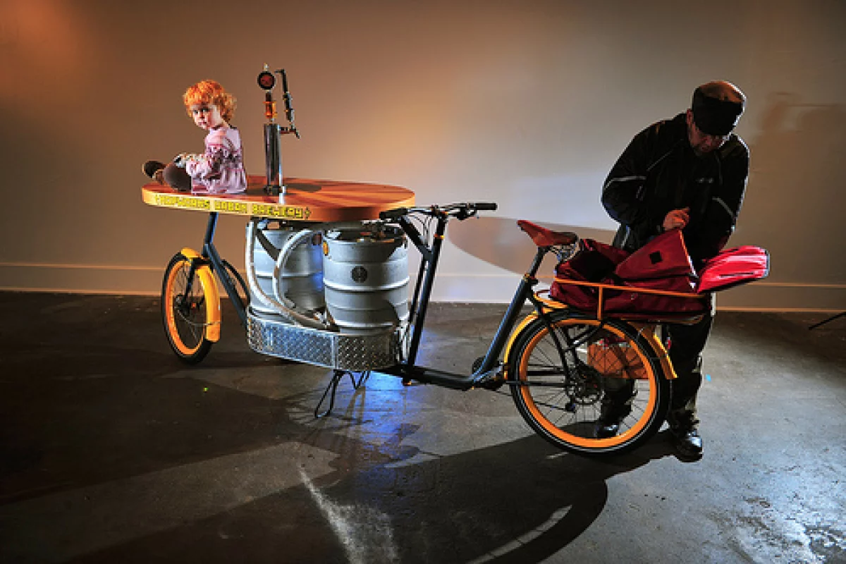 The Beer Bike is a custom-built cargo bicycle that features two beer kegs, a wooden bar with two serving taps, space for pizza boxes, and a solar-powered boom box