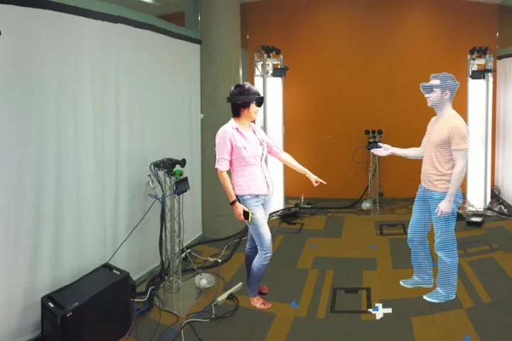 A person on the left wearing a HoloLens interacts with a 3D rendering of another person via holoportation on the right