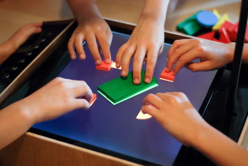 A child arranges corresponding colored blocks over the shapes. With the pieces in place, the "record" button is pressed, and the camera above, which points down at the screen, takes a snapshot