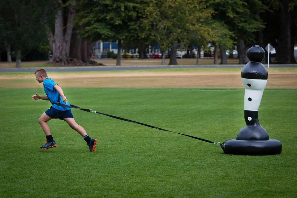 The Shadowman Junior sits on top of a separate tire-like base that's dragged across the turf by a coach or another player using an attached strap/harness, as players are trying to tackle it