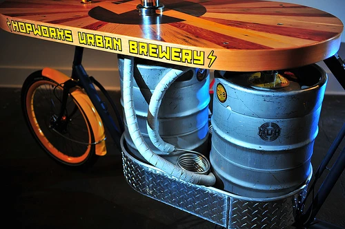 The Beer Bike is a custom-built cargo bicycle that features two beer kegs, a wooden bar with two serving taps, space for pizza boxes, and a solar-powered boom box