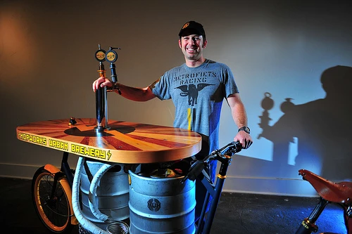 The Beer Bike is a custom-built cargo bicycle that features two beer kegs, a wooden bar with two serving taps, space for pizza boxes, and a solar-powered boom box