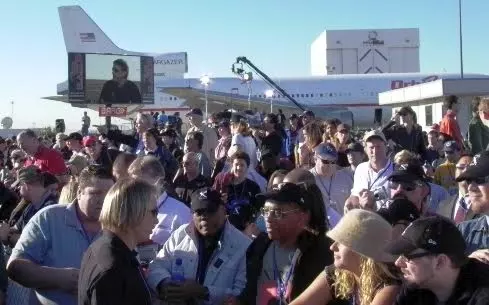 While waiting for Spaceship One to land, Dezso shares details with musicians Quincy Jones and Herbie Hancock and other spectators. Erik Lindbergh appears on the large video screen.