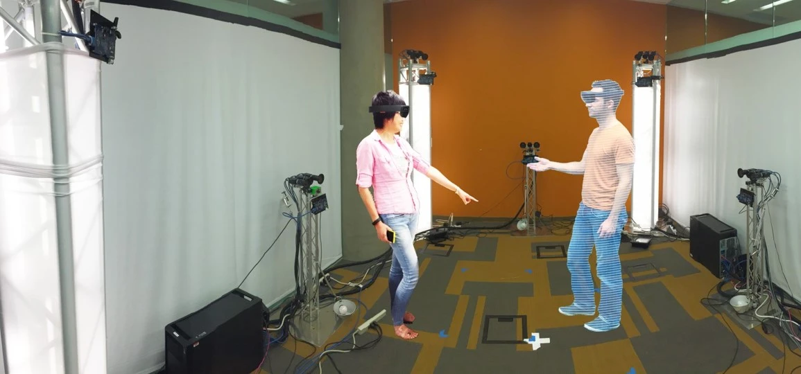 A person on the left wearing a HoloLens interacts with a 3D rendering of another person via holoportation on the right