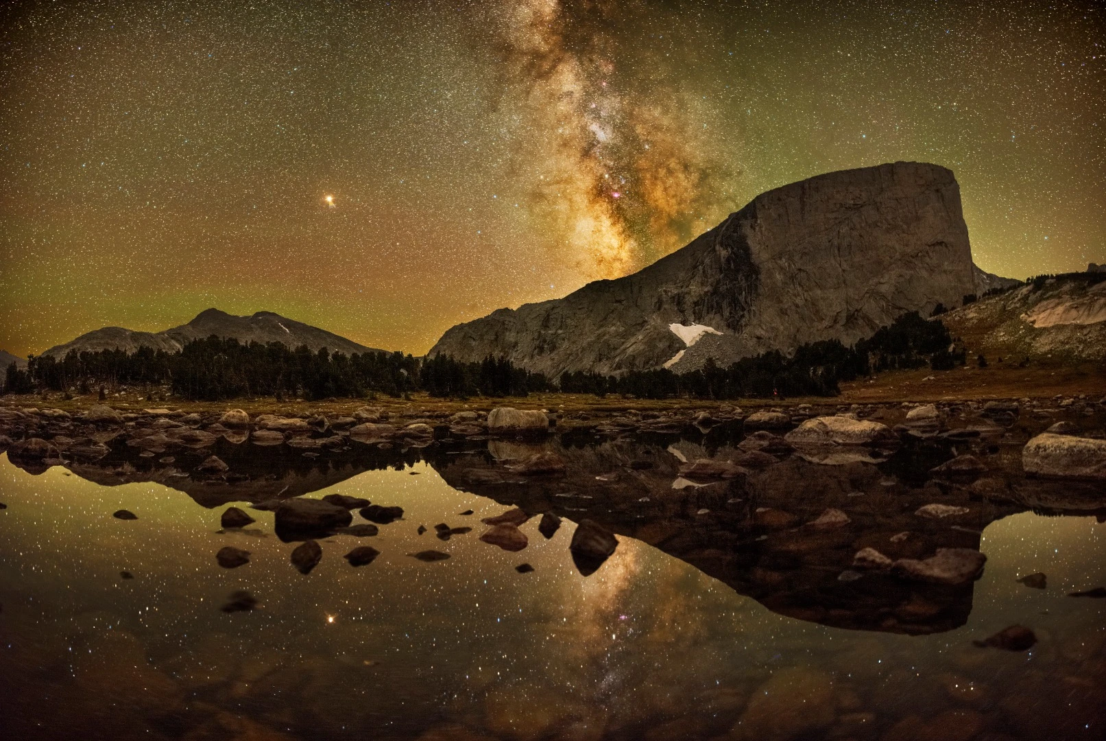 Mount Hooker sits 15 miles past alpine lakes and mountain passes in the Wind River Mountain Range in Wyoming. After the sun set the photographer ventured across the lake near camp. The lake was shallow; at its far end, the low water revealed stones of various sizes. Following the narrow tunnel of the headlamp's beam, the photographer leapt from rock to stone across the water. The sky was clear, the glorious Milky Way was looming over the mountains and the stars shone bright and everything was reflected beautifully on the foreground. The photographer sat on a rock about 20 feet into the lake and started to shoot. Engulfed in dark beauty the photographer realized that astrophotography is primarily about the experience, with the beautiful photo just an extra. Pinedale, Wyoming, USA, 3 September 2018