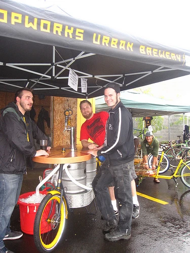 The Beer Bike is a custom-built cargo bicycle that features two beer kegs, a wooden bar with two serving taps, space for pizza boxes, and a solar-powered boom box