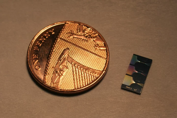 The photonic chip next to a UK penny. The chip contains micrometer and sub-micrometer features and guide light using a network of waveguides. The output of this network can be seen on the surface of the chip.