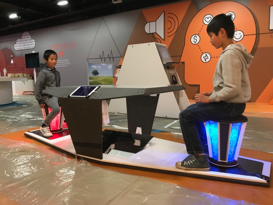 Two players control their movements in a game of electronic ping-pong by swinging their pelvis on a couple of the stools