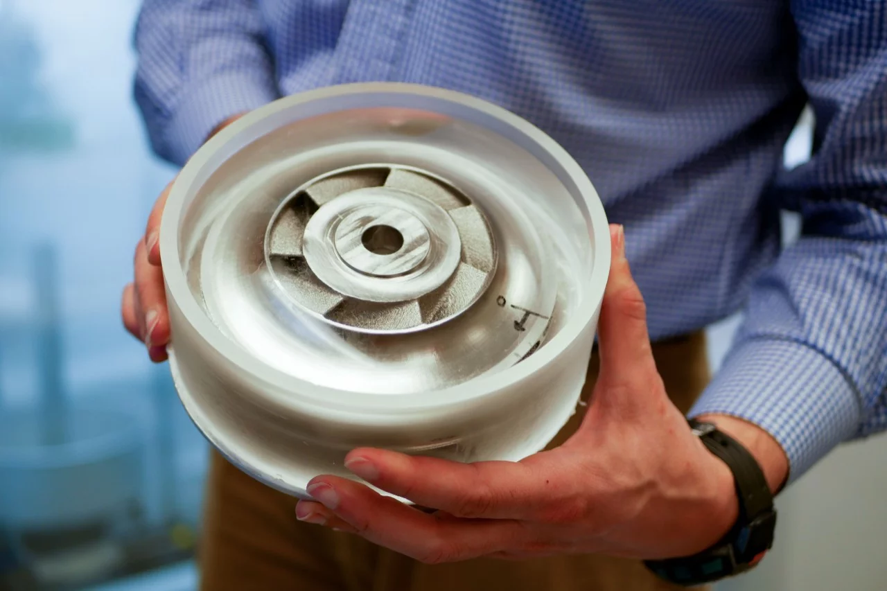 A round 3D-printed metal part, surrounded by ultra-clear ice within a plastic cylinder