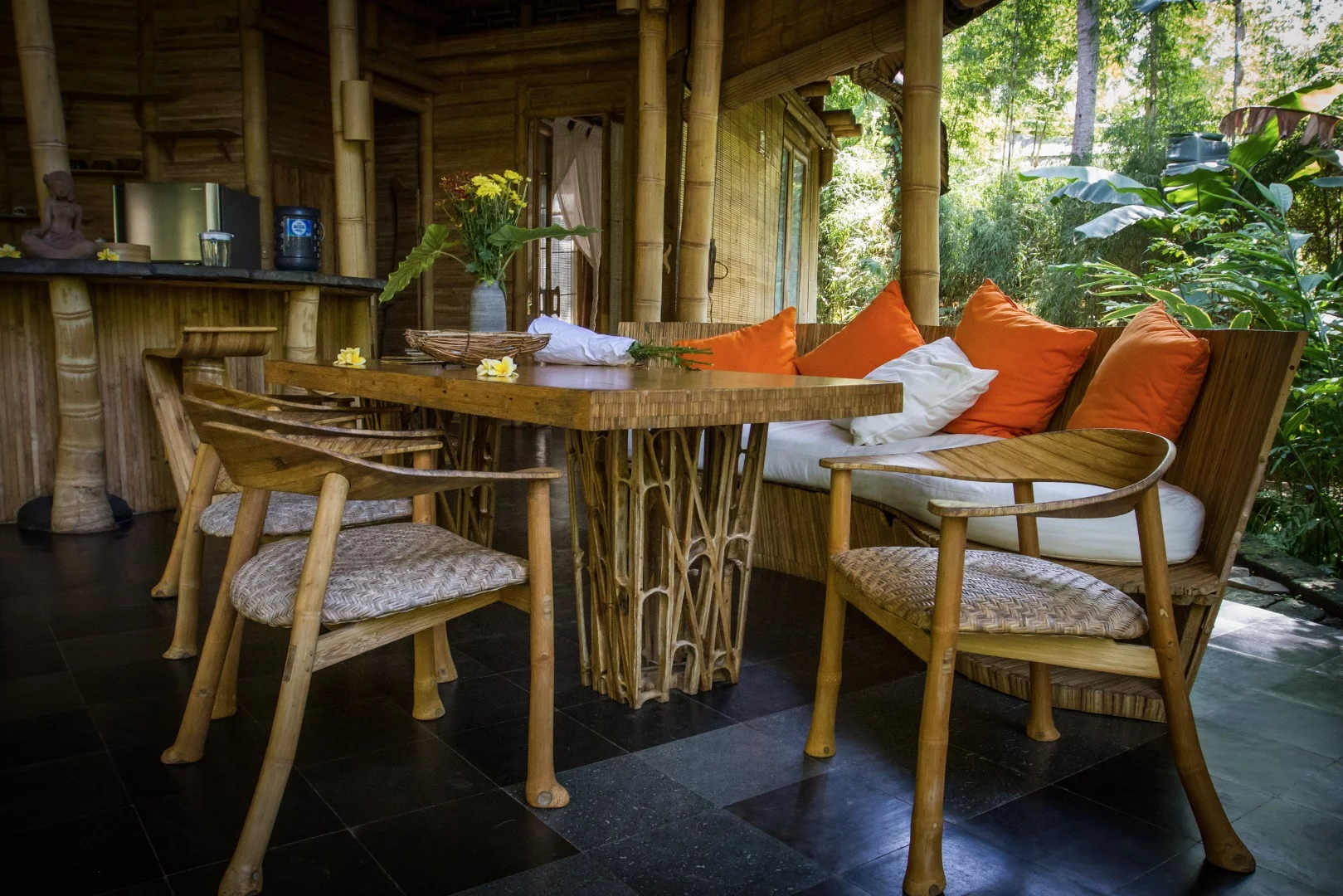 A bamboo table sits surrounded by bespoke chairs, bench seating and lush green foliage
