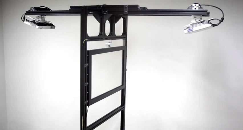 The system is housed within a T-shaped frame that also incorporates two overhead-mounted projectors, which project visuals onto either side of the screen