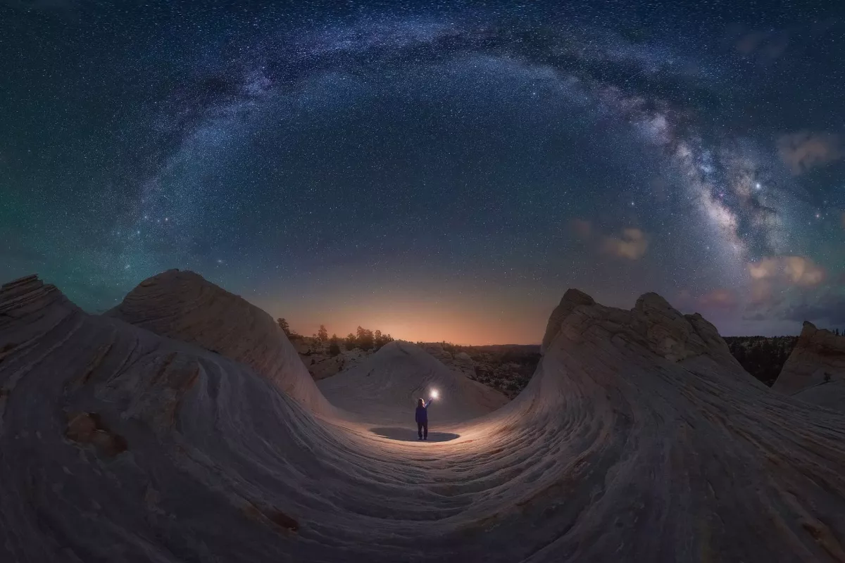 Good Night Utah, shot in Kanab, Utah, USA. Waves of rock mimic the arch of the Milky Way overhead, forming a makeshift circle. The Sun begins to peek over the horizon, the same color as the light cast by the figure in the center.