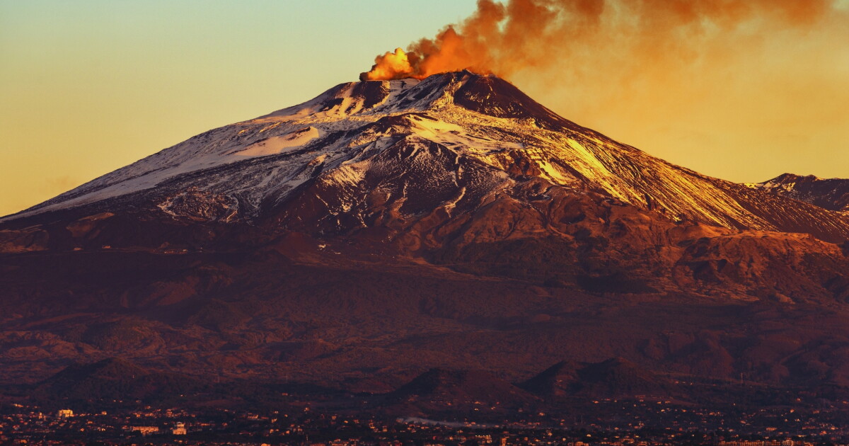 Büyük, karla kaplı bir stratovolkan olan Etna Dağı, sıcak bir günbatımı gökyüzüne karşı turuncu ve sarı lavdan oluşan bir duman püskürtür.