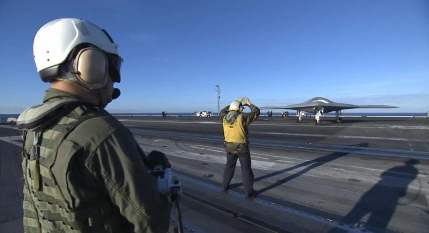 An operator (foreground) uses a wireless arm-mounted control display unit to command the X-47B UAV as it taxis aboard the USS Truman aircraft carrier