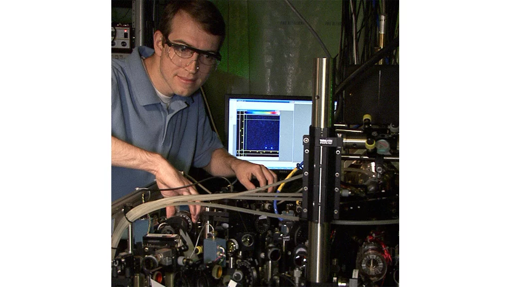 NIST postdoctoral researcher David Hanneke at the laser table used to demonstrate the first universal programmable processor. The monitor displays a colorized image of the two beryllium ions that hold information in the processor (Photo: J. Burrus/NIST)