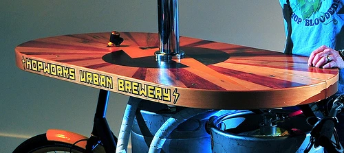 The Beer Bike is a custom-built cargo bicycle that features two beer kegs, a wooden bar with two serving taps, space for pizza boxes, and a solar-powered boom box