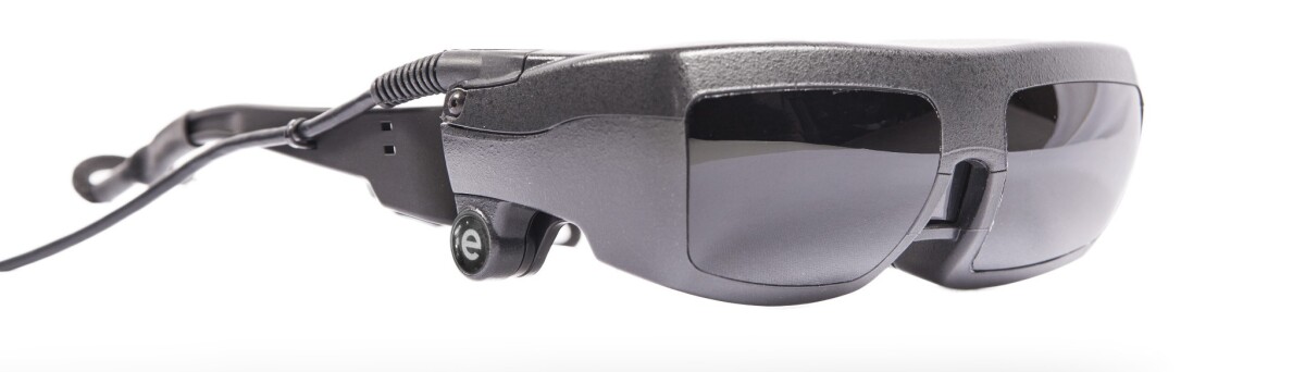 The eSight system consists of a frame that houses the user's prescription lenses, over top of which lies a headset containing an HD video camera and two LCD screens, along with a hand-held hard-wired control unit