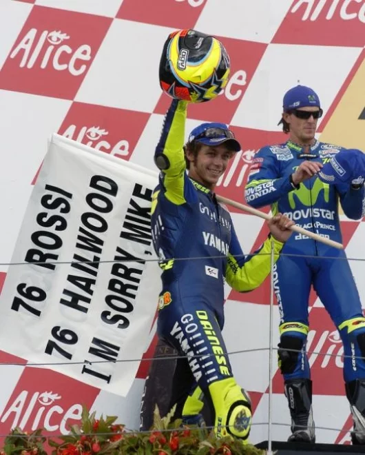 Vale climbs the podium with his flag while Sete Gibernau retreats behind his shades to wonder what he has to do to beat Rossi