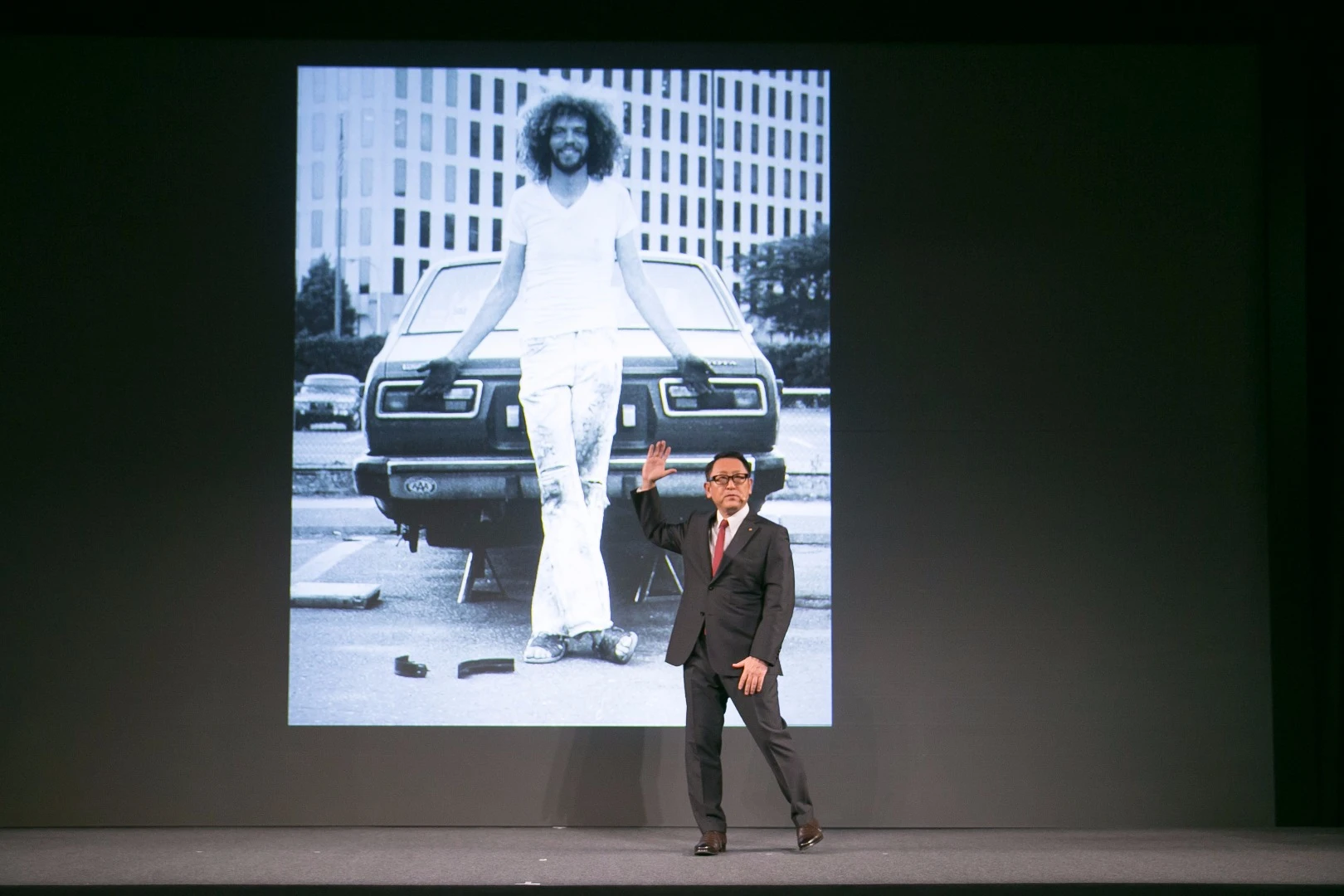 Akio Toyoda, President of Toyota Motor Corporation show a slide of a younger Gill Pratt in front of his Toyota Corolla