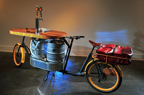 The Beer Bike is a custom-built cargo bicycle that features two beer kegs, a wooden bar with two serving taps, space for pizza boxes, and a solar-powered boom box