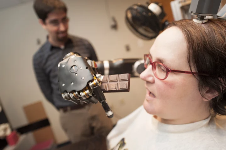 Quadriplegic Jan Scheuermann prepares to take a bite out of a chocolate bar she is guiding into her mouth with a thought-controlled robot arm while research assistant Brian Wodlinger, Ph.D., watches on (Photo: UPMC)
