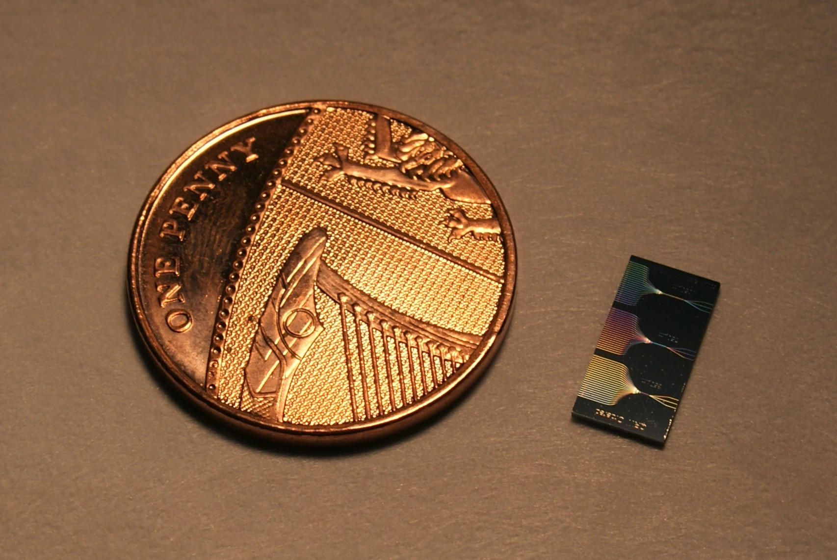 The photonic chip next to a UK penny. The chip contains micrometer and sub-micrometer features and guide light using a network of waveguides. The output of this network can be seen on the surface of the chip.
