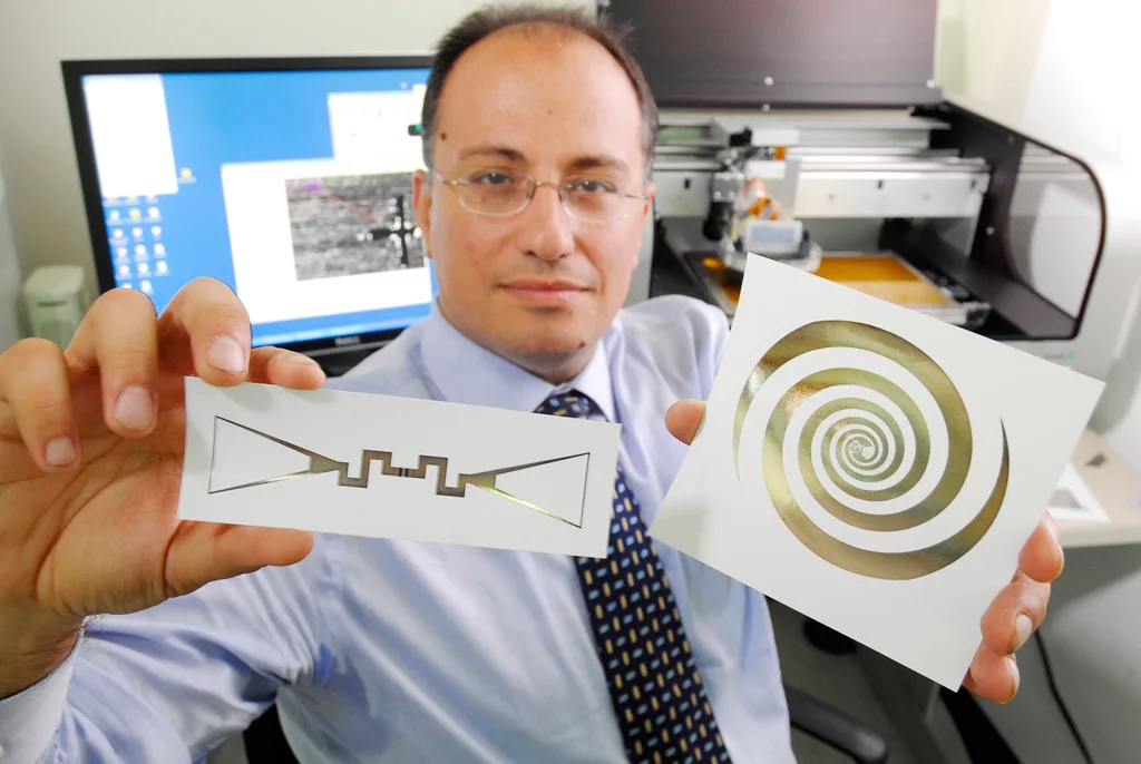 Manos Tentzeris holds a sensor (left) and an ultra-broadband spiral antenna for wearable energy-scavenging applications that were both printed on paper using inkjet technology (Image: Gary Meek)