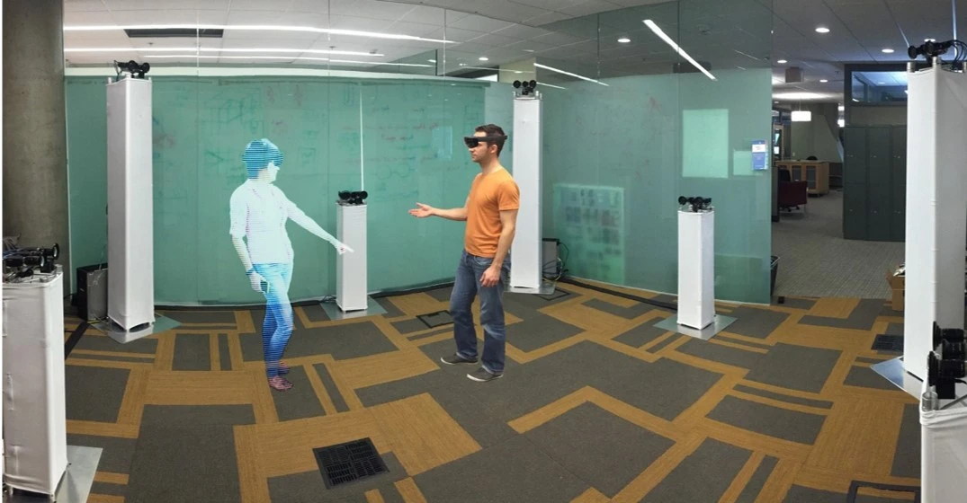 A person on the right wearing a HoloLens interacts with a 3D rendering of another person via holoportation on the left