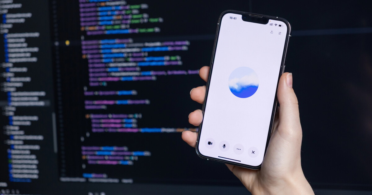 Bir kişinin eli, bilgisayar ekranında kodla dolu karanlık bir arka plan üzerine yerleştirilmiş bulutlu gökyüzü fotoğrafını gösteren bir iPhone'u tutar.