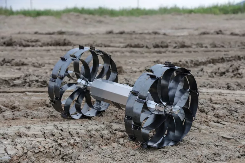 Dispositivo robótico con dos ruedas formadas por palas metálicas curvadas, unido por un eje central, colocado sobre un terreno de tierra arada en un entorno agrícola o experimental.