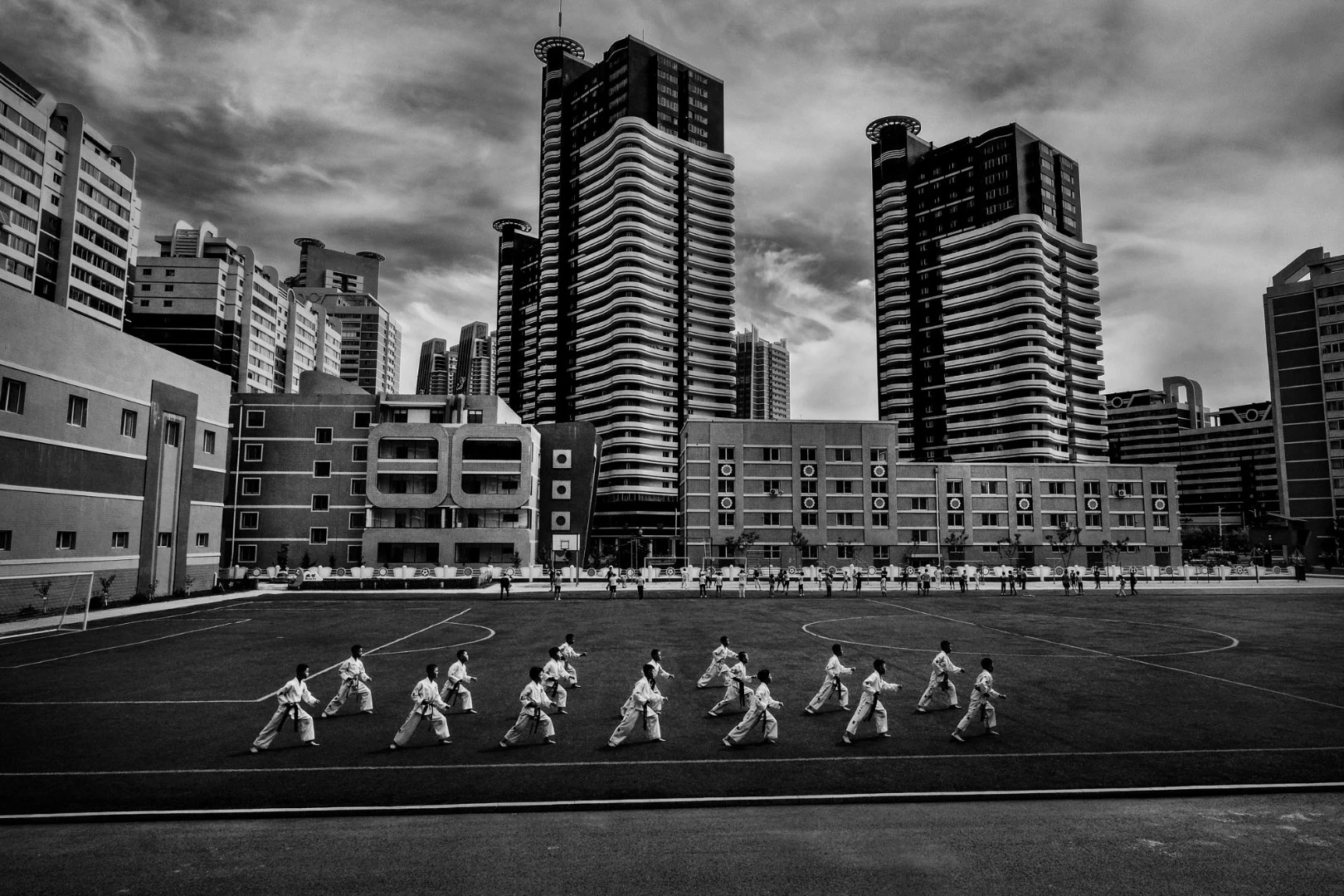 Pyongyang, North Korea. On the football field adjacent to Mirae Primary School, with the typical skyscrapers of Pyongyang in the background, children practice the synchronized execution of a tul, a series of offensive and defensive techniques against one or several virtual adversaries
