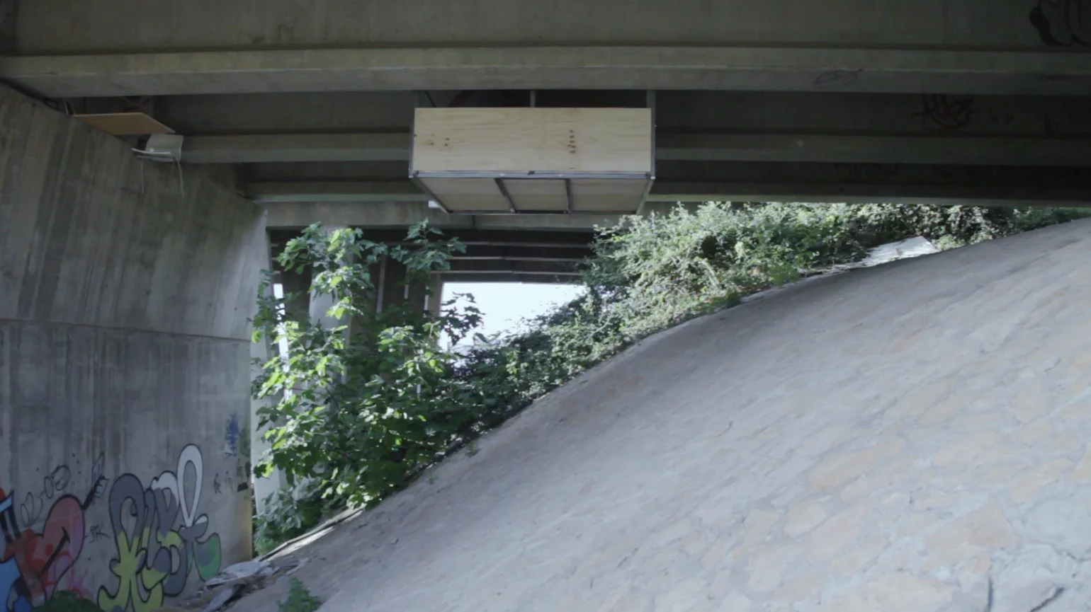 A visitor walks up the banked area and enters the plywood-lined metal shelter, then uses an available hand crank to move it along the bridge's underside to where a bench, seat, and some shelving are waiting