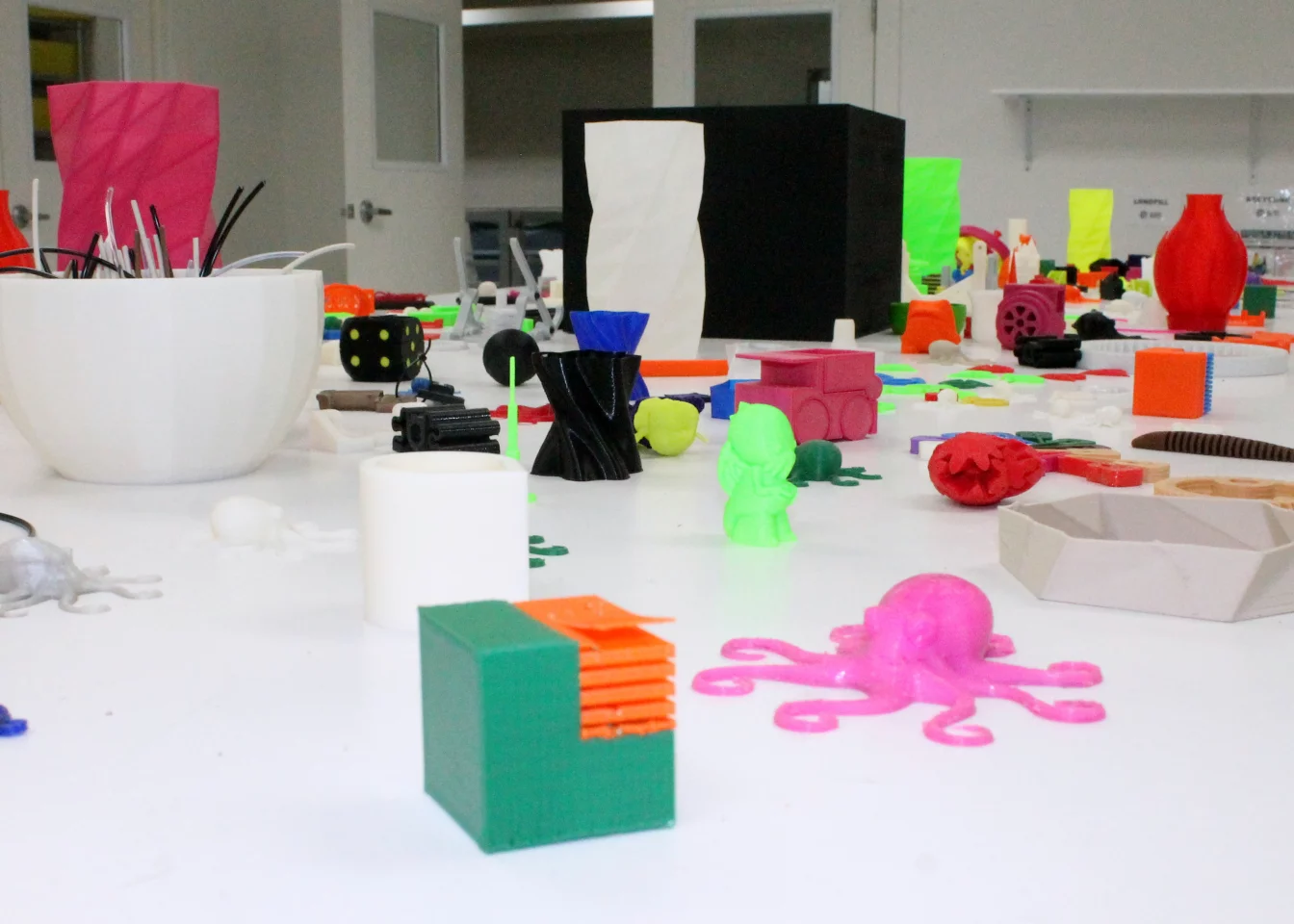 The printer factory features a table of different printed examples, toys, gears, and tools illustrating LulzBot filament types, print quality, and practicality