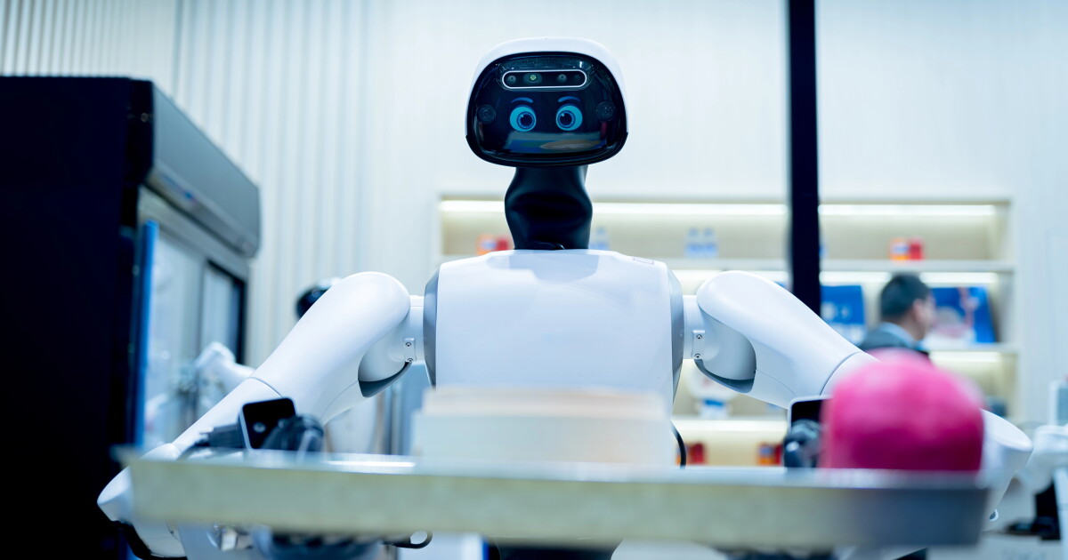 Parlak aydınlatmalı, arka planı bulanık bir odada, pembe bir nesne taşıyan tepsiye sahip beyaz bir robot konumlandırılmıştır.