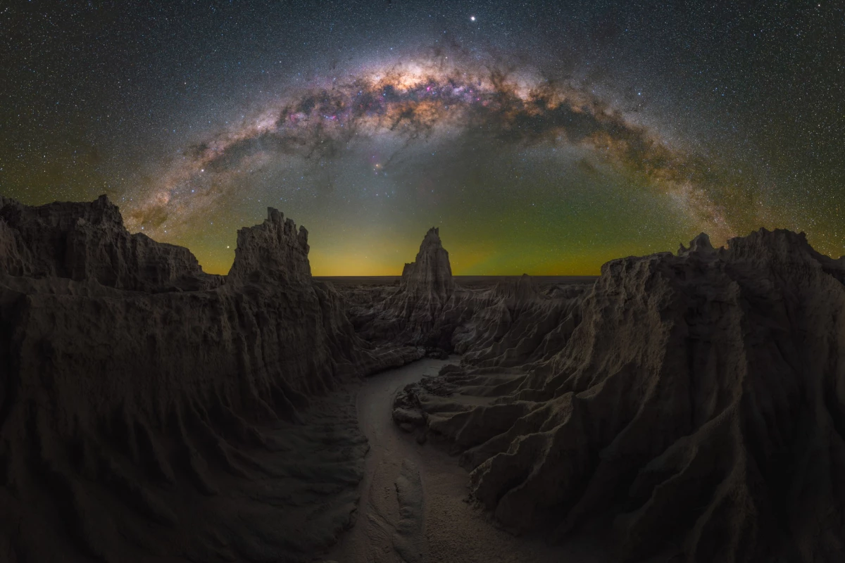 Dragon's Lair, shot in Mungo, New South Wales, Australia. The Milky way arcs over a canyon where jagged cliffs frame a winding path, leading to a central spire. A faint golden glow emanates from the horizon.