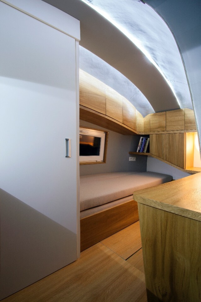 The interior comprises a small seating area and workspace, a bed, toilet and shower area, and a kitchenette