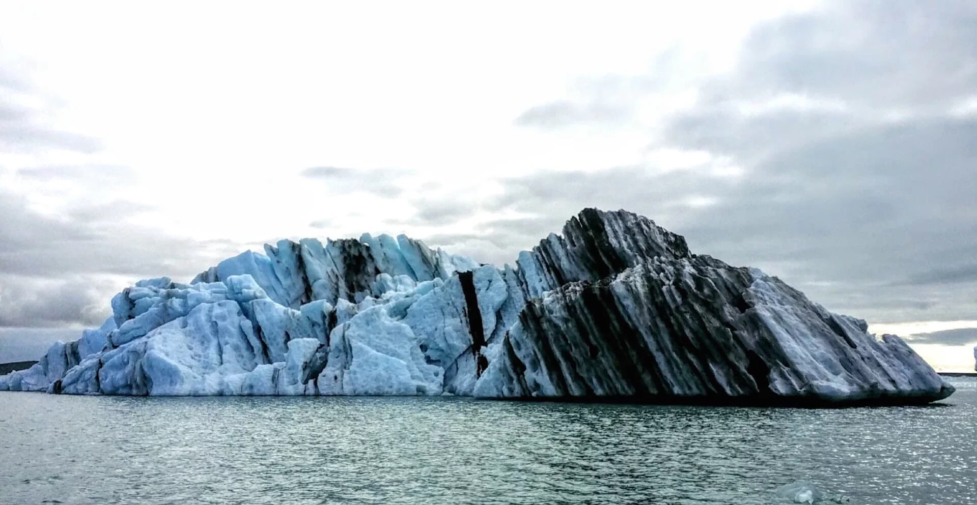The color of an iceberg can tell you a lot. In this berg, the stripes of black represent volcanic eruptions that spewed ash on top of ice over time. The blueish tint to the ice shows that the iceberg flipped over recently. That's because when the ice first calves from the glacier, it is compacted so tightly that light coming through it takes on a blue hue. After the iceberg flips over, as they do from getting top heavy, the ice eventually turns white as the sun creates tiny cracks in its structure.