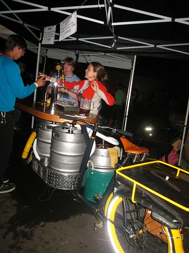 The Beer Bike is a custom-built cargo bicycle that features two beer kegs, a wooden bar with two serving taps, space for pizza boxes, and a solar-powered boom box