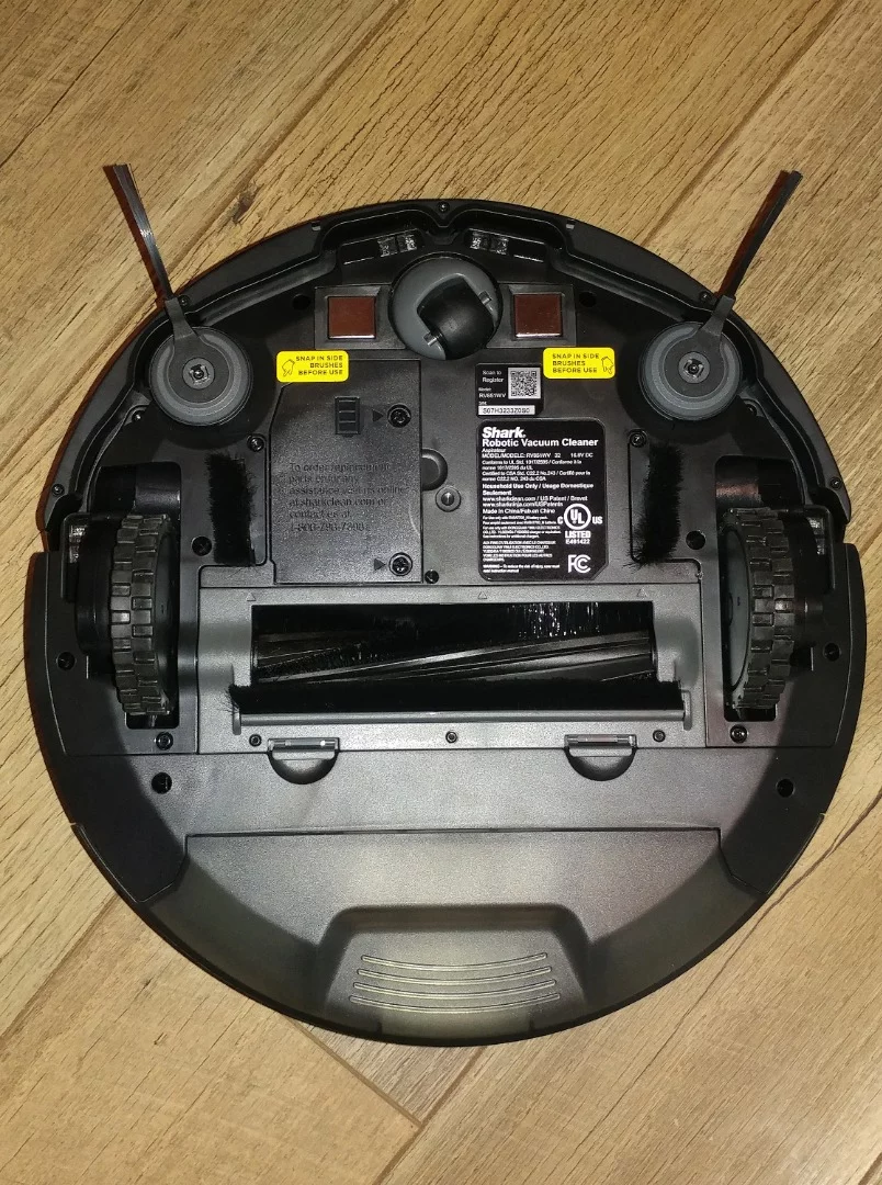 The underside of the Shark Ion vacuum shows the two drive wheels (center edges), the rotating brush and vacuum port (center) between them, and the third wheel for stability (top)