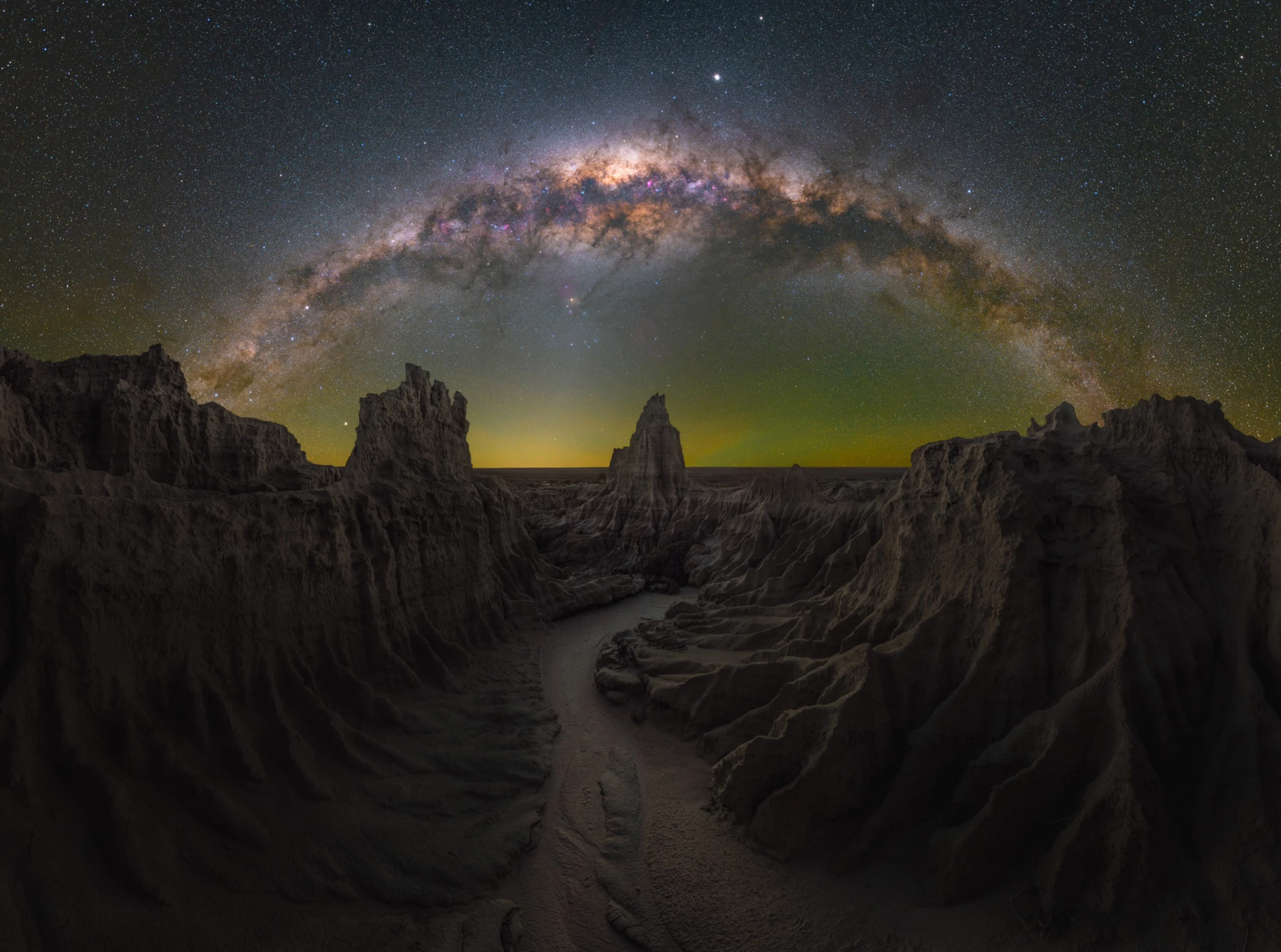 Dragon's Lair, shot in Mungo, New South Wales, Australia. The Milky way arcs over a canyon where jagged cliffs frame a winding path, leading to a central spire. A faint golden glow emanates from the horizon.