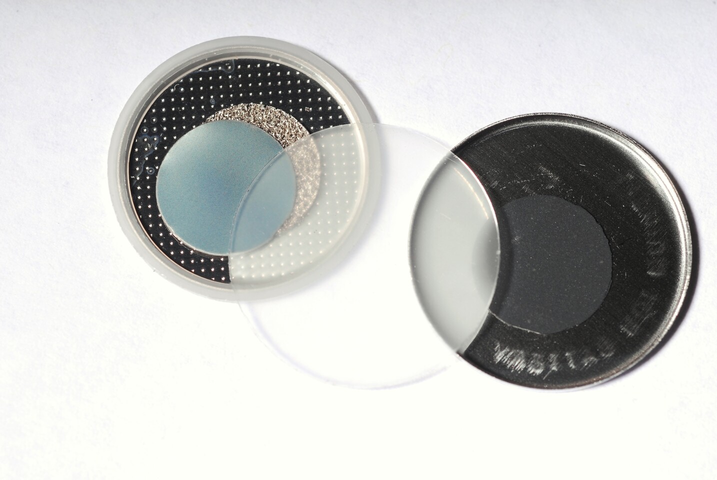 Components of a new self-charging piezoelectric power cell – the clear disc in the center is the piezoelectric film that serves as a charge pump for lithium ions (Credit: Gary Meek)