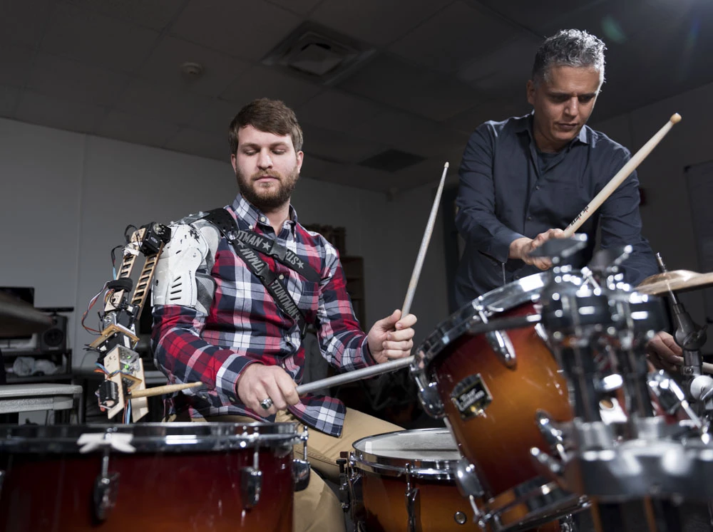 Tyler White (left) with the "third arm" robotic drumming arm accompanied by Professor Gil Weinberg (right)
