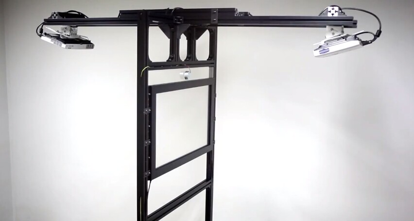 The system is housed within a T-shaped frame that also incorporates two overhead-mounted projectors, which project visuals onto either side of the screen